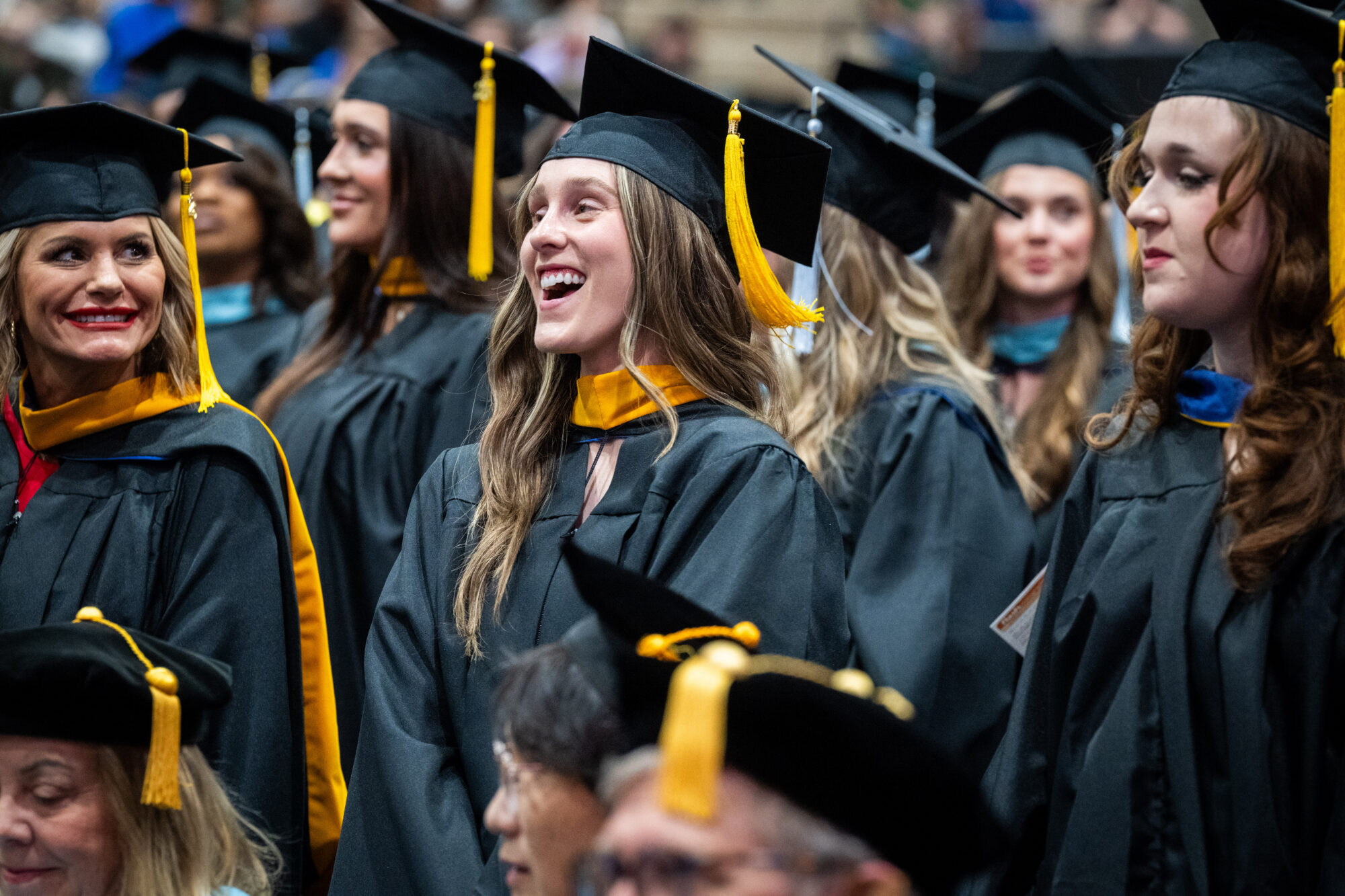 A group of graduates in black caps and gowns, with gold tassels on their caps, smiles happily during the ceremony.