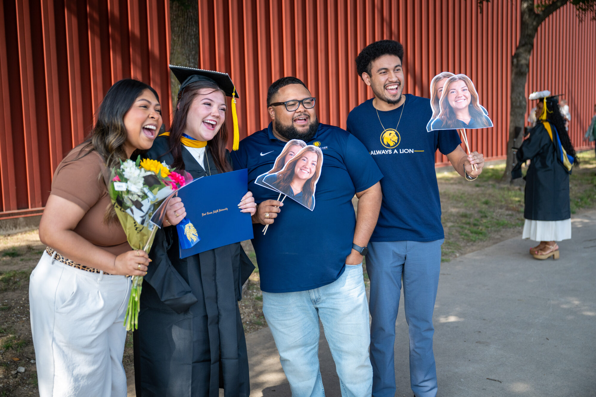 One graduate in a black cap and gown is flanked with three other people. They are all smiling, and two of them are holding cardboard cutouts, on sticks, of the graduate's face. 