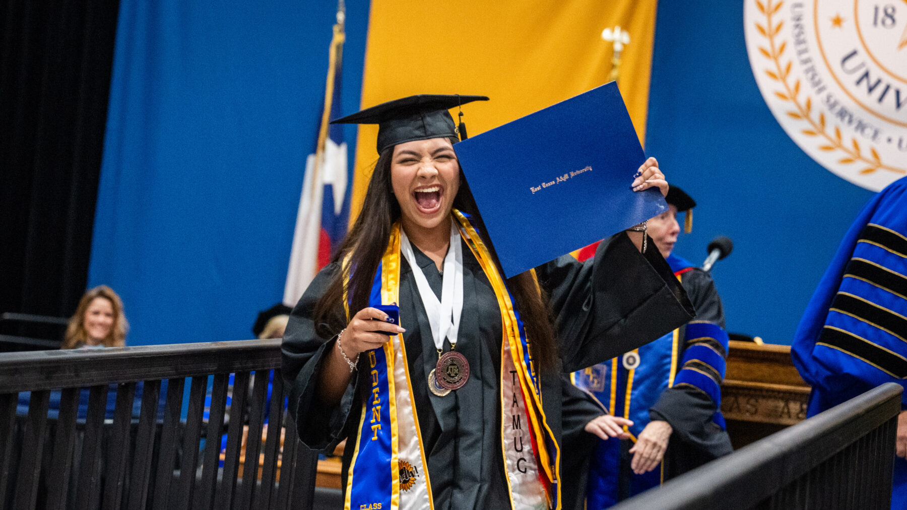 A graduate in black robe and black graduation cap is looking at the camera and has her mouth open as if yelling in celebration. She is holding up her diploma.