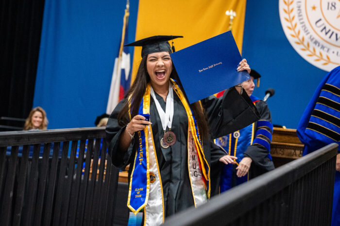 A graduate in black robe and black graduation cap is looking at the camera and has her mouth open as if yelling in celebration. She is holding up her diploma.