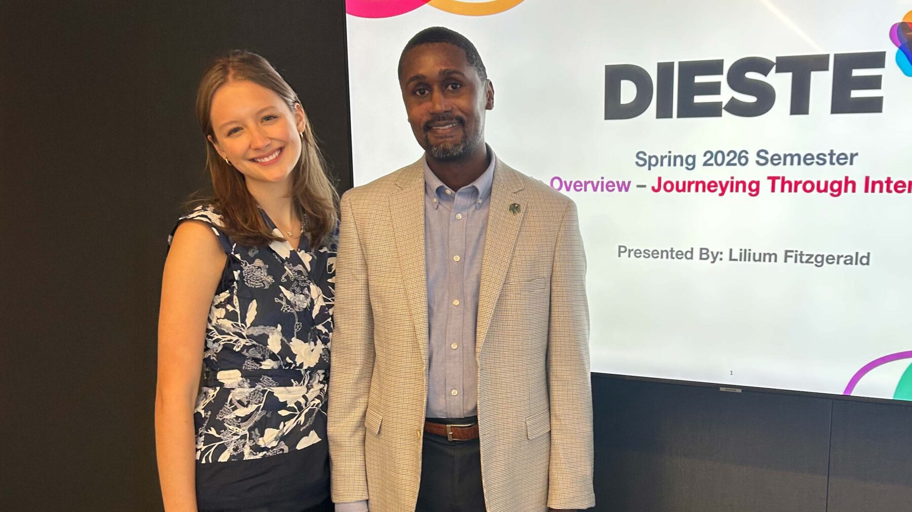 A woman and a man stand side by side in front of a screen that reads Dieste.