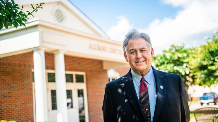 Ted Crim smiles at the camera, dressed in a dark suit and red tie, in front of a red brick building with white gables and white columns.