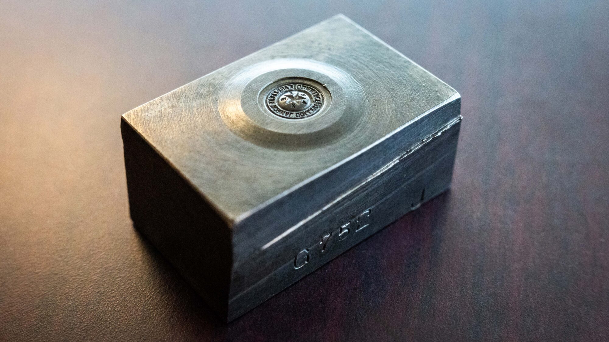 A rectangular artifact sits on a wood table top surface. The artifact, known as a seal die, appears to be made of  tarnished bronze metal.
