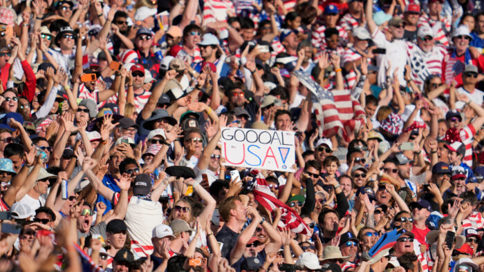 A large number of fans in the crowd of a soccer game. A person is holding a sign the reads "GOOOOAL USA."