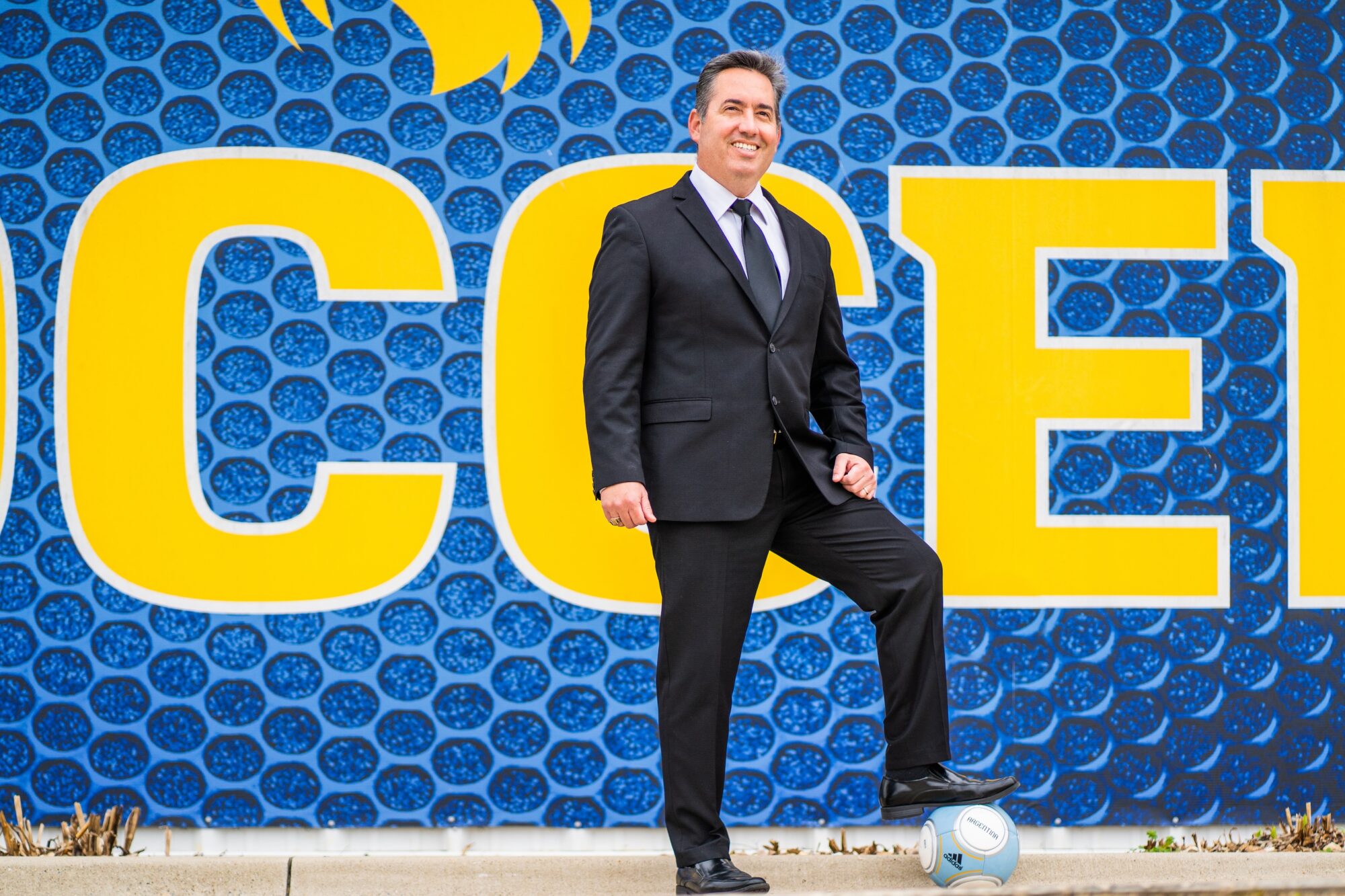 A person in a suit and tie posing with his foot on a soccer ball.