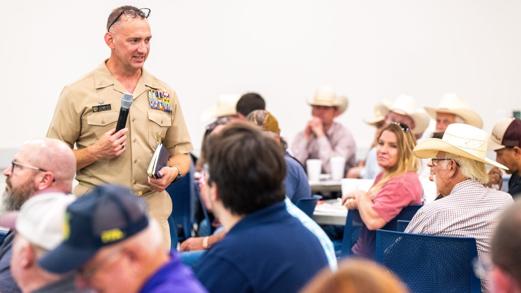 A person in a khaki military uniform speaking to a group with a microphone.