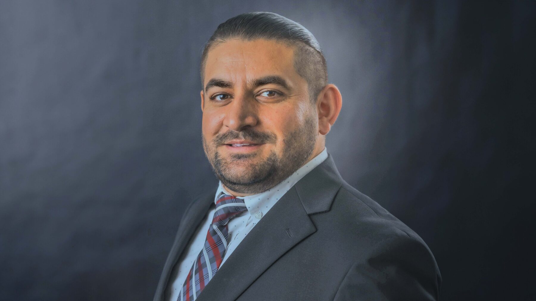 Mashal Mustafa looks at the camera in a professional headshot. He is wearing a grey suit and blue shirt with a tie. He is smiling at the camera, and the background behind him is grey.