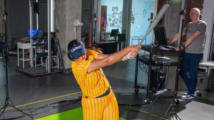 A woman swings a softball bat, with a man watching in the background.