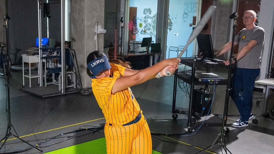 A woman swings a softball bat, with a man watching in the background.