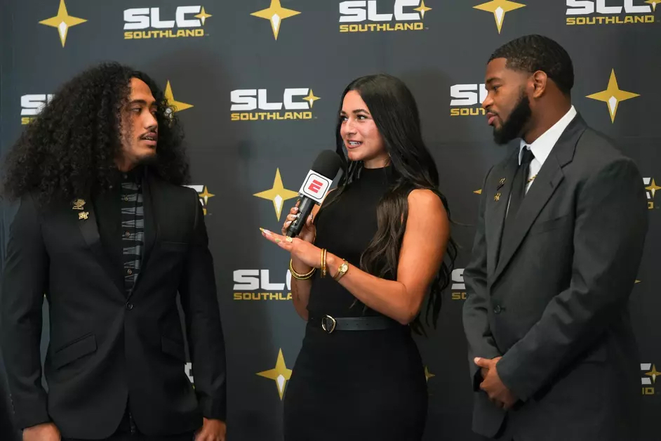 An interviewer speaking with two guests during a football media conference.
