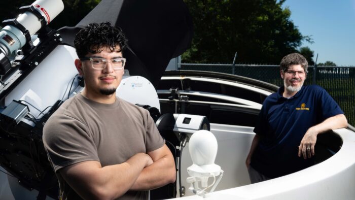 Two men stand outside beside a telescope.