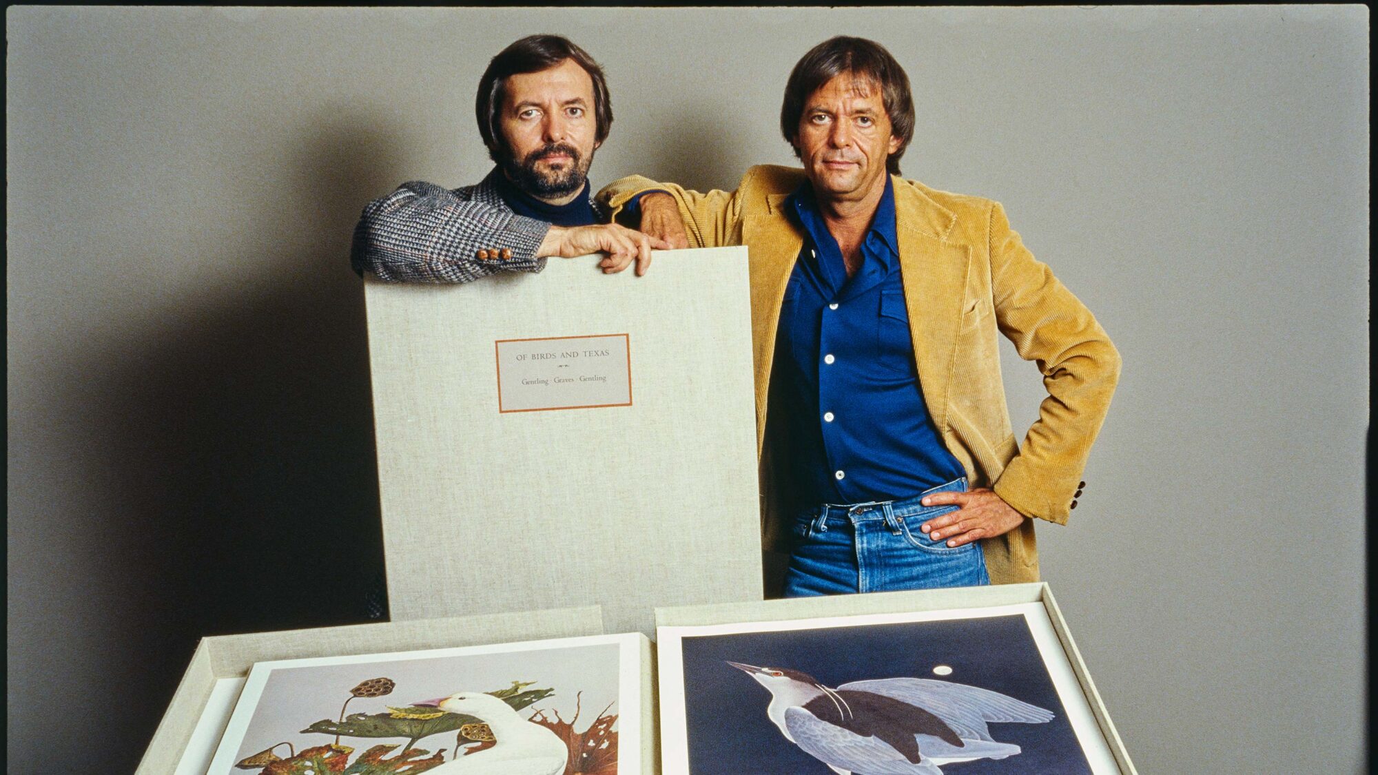 Two men stand together in front of a grey background. The artist on the left is resting his forearm on a large white book, and two bird paintings are displayed below them.