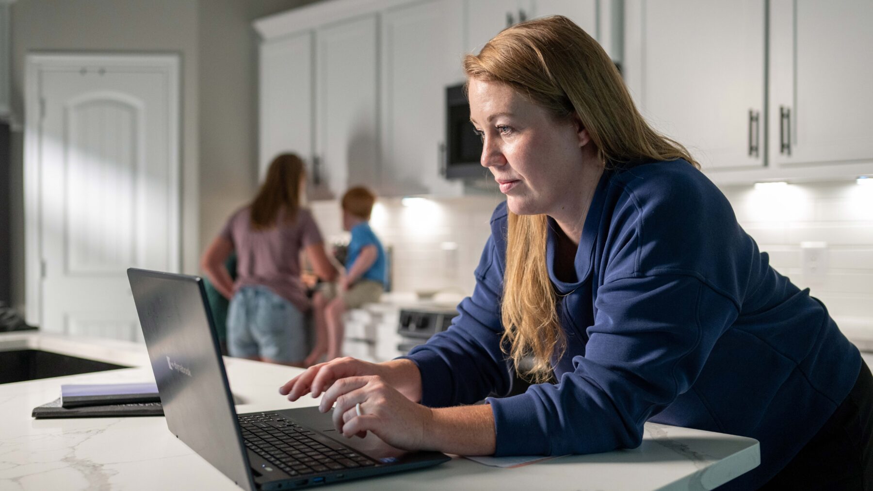 A woman leans over a laptop while kids play in the background.