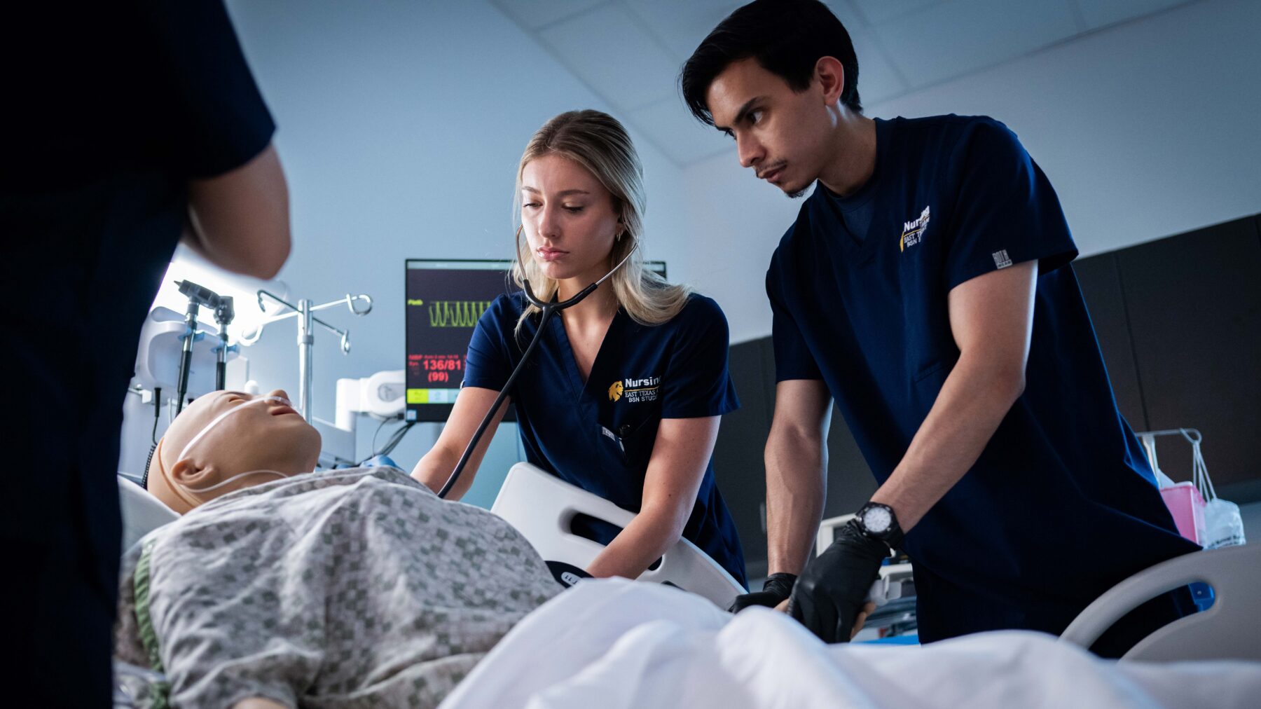 A man and a woman in scrubs look over a mannequin lying in a bed.