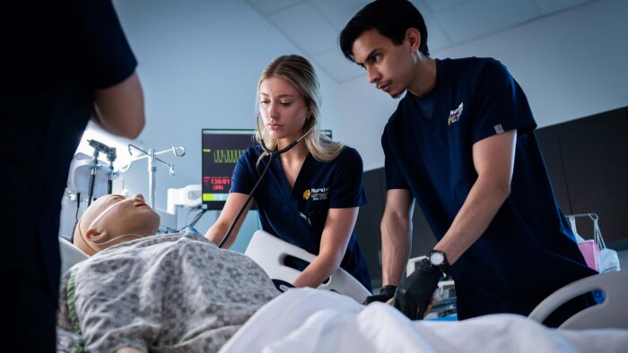 A man and a woman in scrubs look over a mannequin lying in a bed.