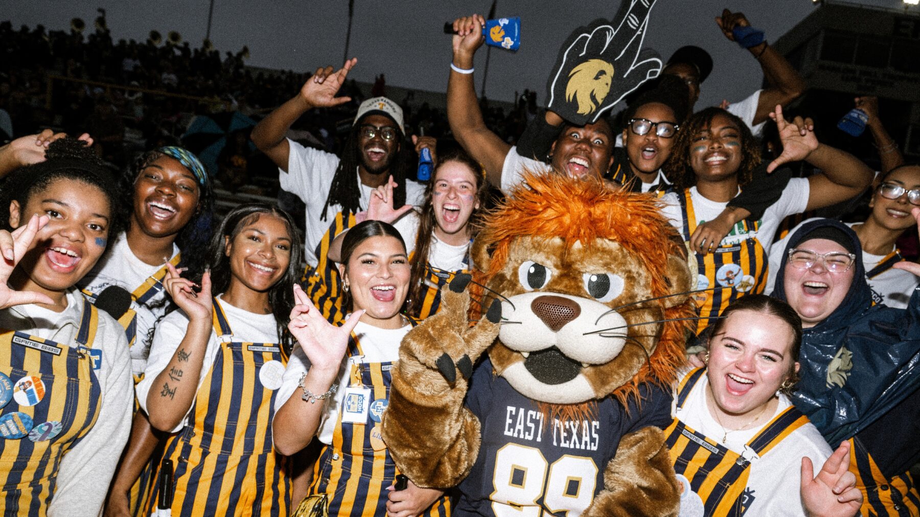 A group of college students and a person in a lion mascot costume in the stands at a football game.