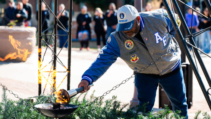 A person in a letterman jacket adorned with U.S. air force regalia uses a gas torch to light a flame.
